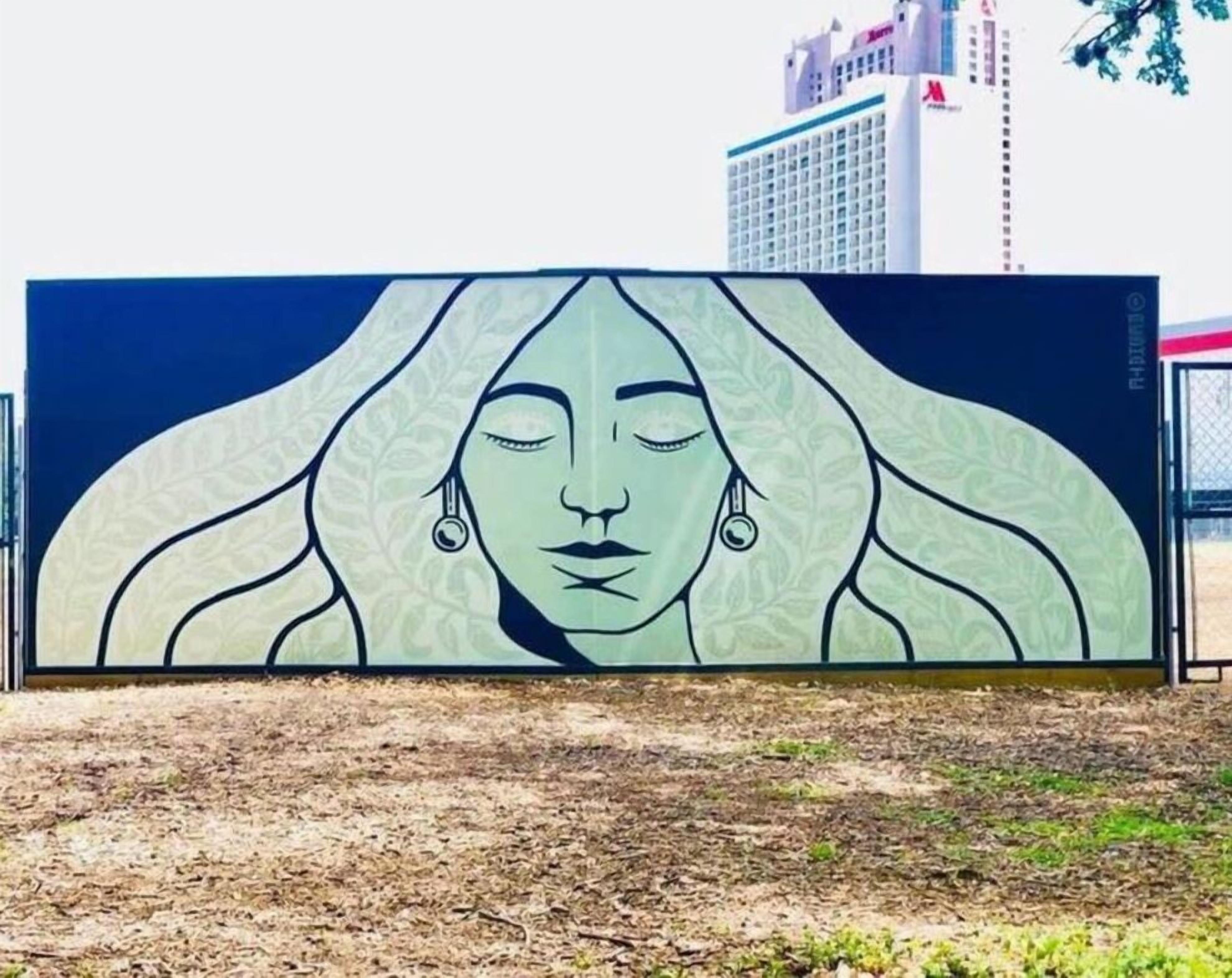 A large outdoor mural with a woman's face, a building in the background, and a fence.