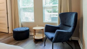 A room with a dark blue armchair, a round wooden table, and a matching ottoman. There are two windows with blinds, and a door is visible to the left. - Charming Studio W/deck & Garden (San Antonio)