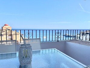 A balcony with a glass table, a glass vase with a drink, and a view of the sea and buildings.