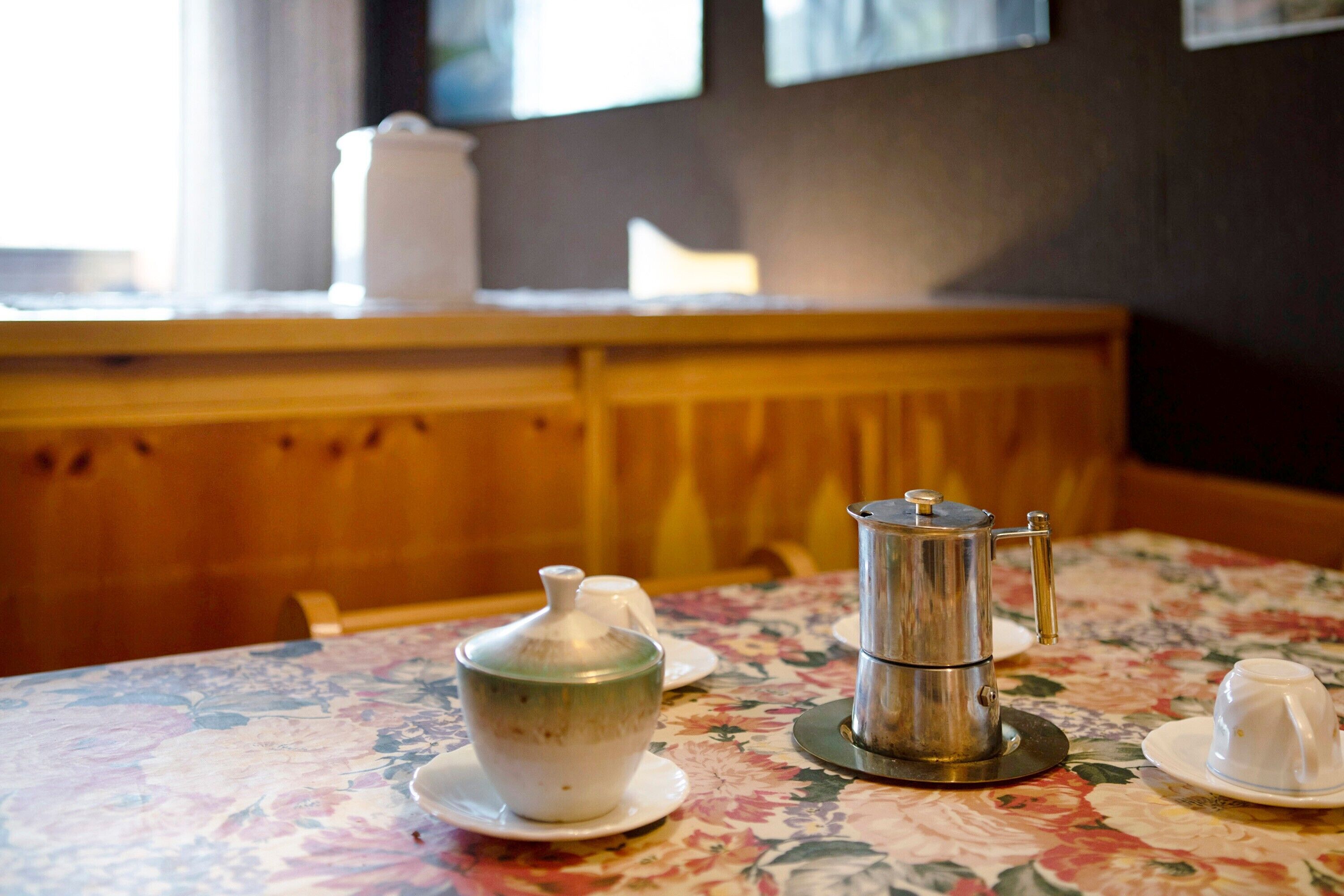 Una mesa con mantel floreado, una cafetera, un hervidor y una taza en una habitación con paneles de madera y un ventanal.