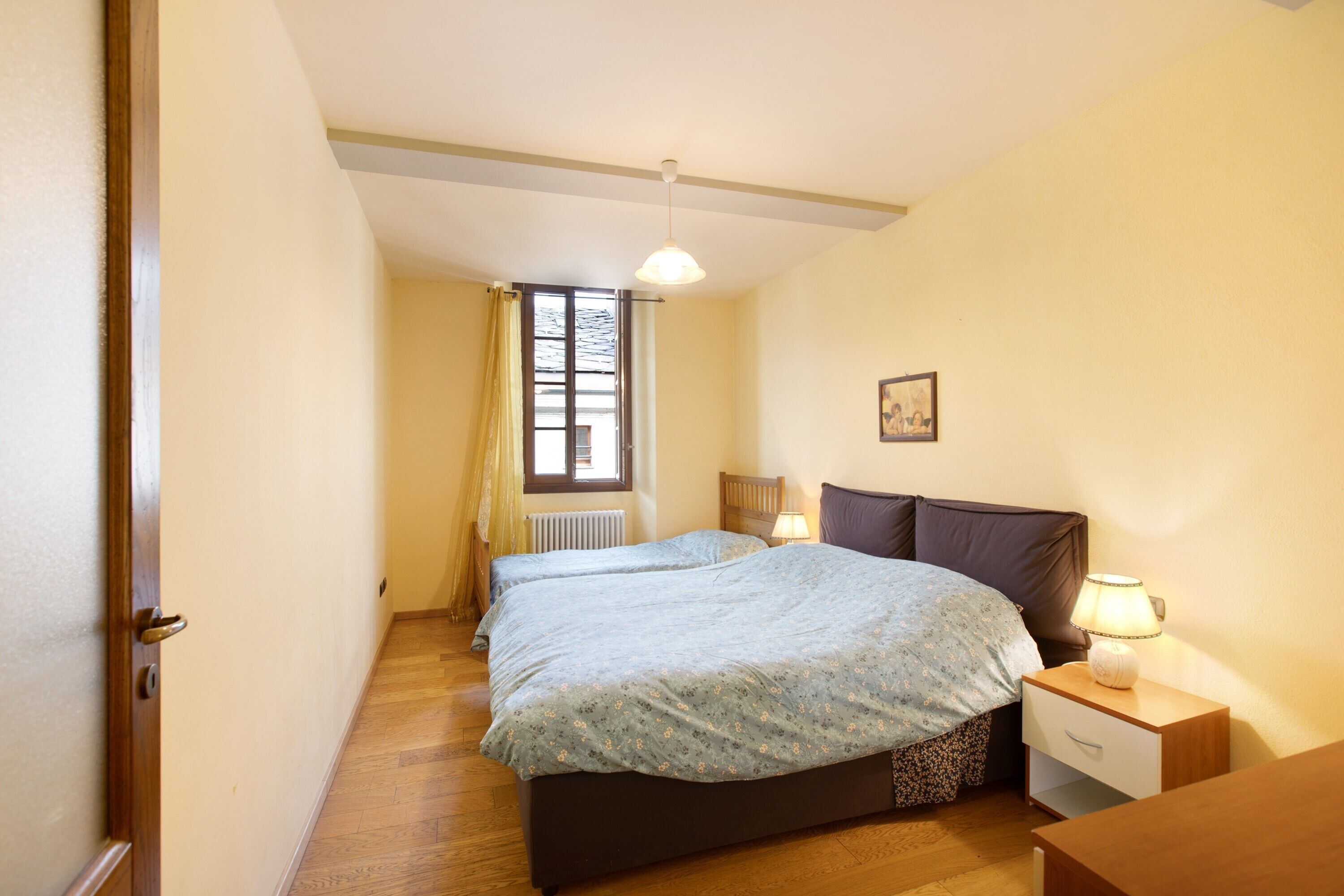 A bedroom with two beds, a nightstand, a lamp, a window with curtains, and a radiator.