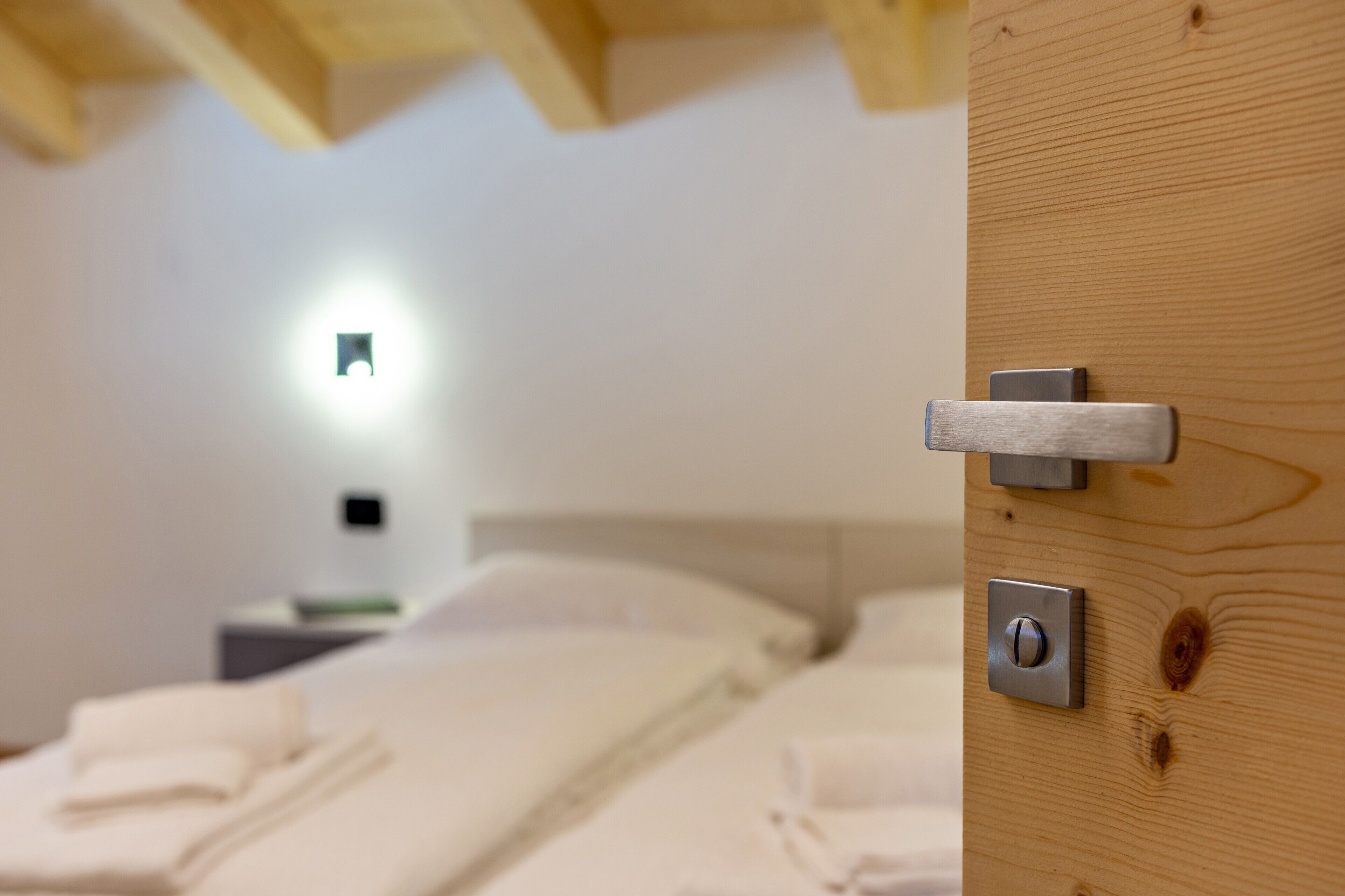 Habitación de hotel con puerta de madera, cama con ropa de cama blanca, mesita de noche y lámpara de pared.