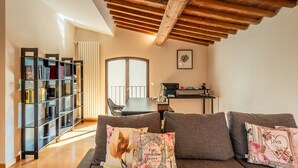 Una sala de estar con techo de madera, una estantería, un sofá con cojines estampados y una zona de comedor con mesa y sillas.
