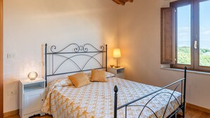 Un dormitorio con estructura de cama metálica, una mesita de noche con lámpara, un ventanal con vistas y una mesita de cabecera con lámpara.