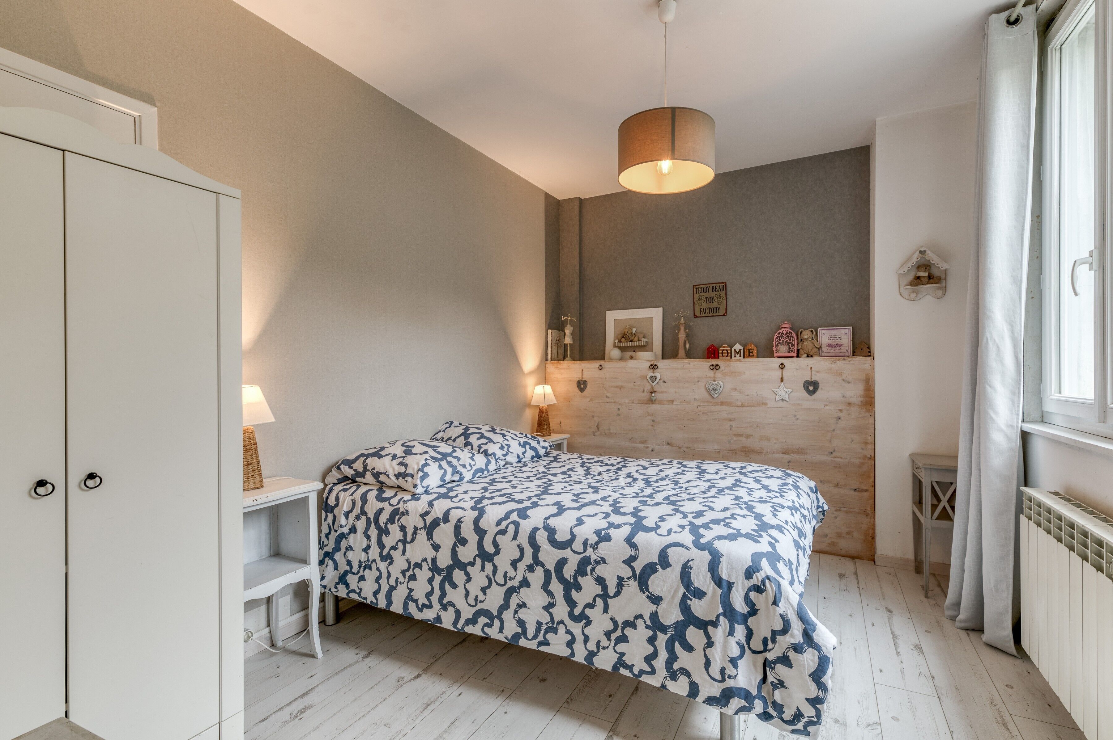 Une chambre à coucher comprenant un lit, des tables de chevet, une armoire, un luminaire suspendu, une fenêtre avec des rideaux et un mur décoré de différents objets.