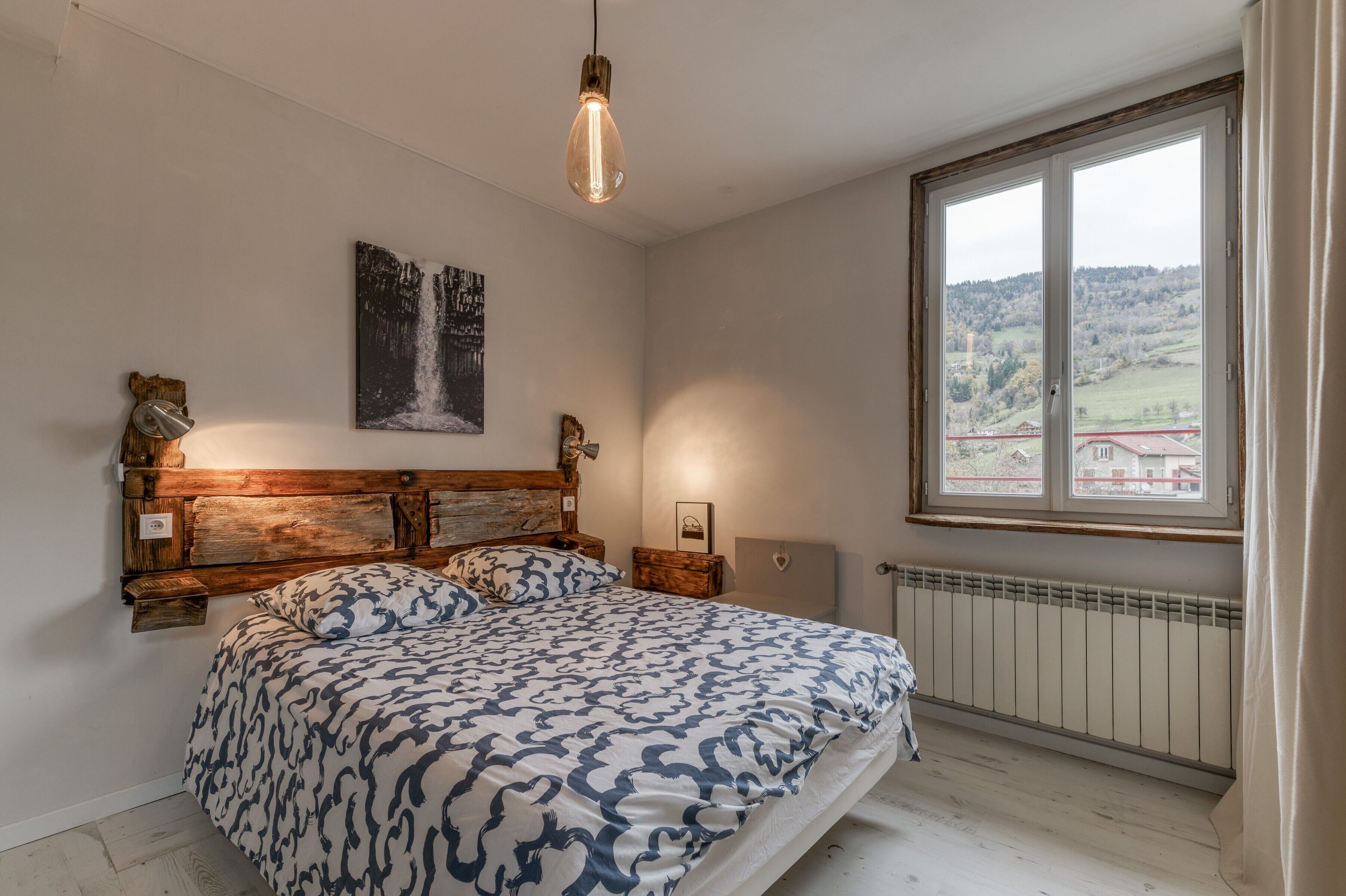 A bedroom with a bed, a wooden headboard, a window with a view, and a hanging light fixture.