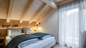 Ein Schlafzimmer mit Holzboden, einem Bett mit Kissen, einem Nachttisch, einer Wandlampe und einem Fenster mit Vorhängen.