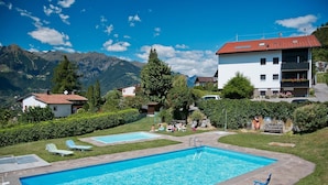 A pool area with two pools, a building with a red roof, and mountains in the background. - Studio Apartment 'Spiess' with Mountain View, Shared Pool and Wi-Fi (Merano)