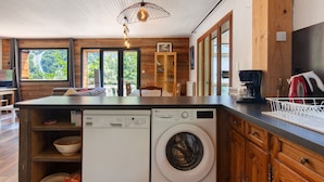 Une cuisine compacte dotée d’un lave-linge et d’un sèche-linge intégrés, de meubles en bois et de grandes fenêtres offrant une vue sur l’extérieur.