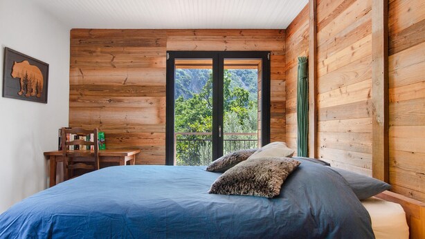 Une chambre à coucher avec un grand lit, un mur en bois, une fenêtre donnant sur des arbres et une bibliothèque.