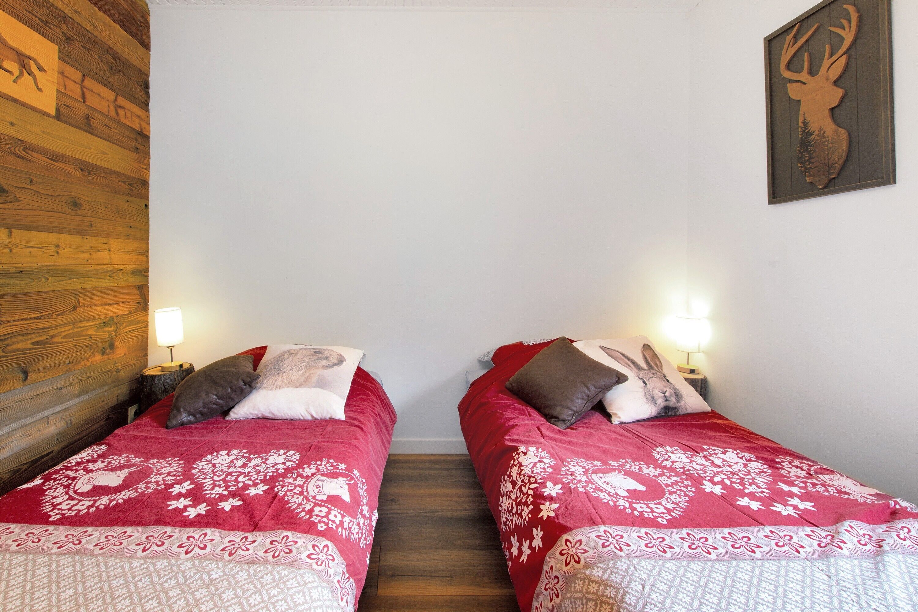 Een kamer met twee bedden, elk voorzien van een rode sprei en decoratieve kussens, een houten wand, en een decoratie van een hertenhoofd aan de muur.