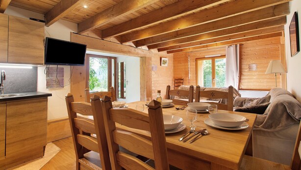 Een houten interieur met een eettafel gedekt voor vier personen, houten stoelen, een aan de muur bevestigde televisie en een raam met uitzicht op groen.