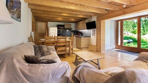Een gezellige woonkamer met houten plafond, een eethoek met tafel en stoelen, een keuken met moderne apparatuur en een schuifpui naar een tuin.