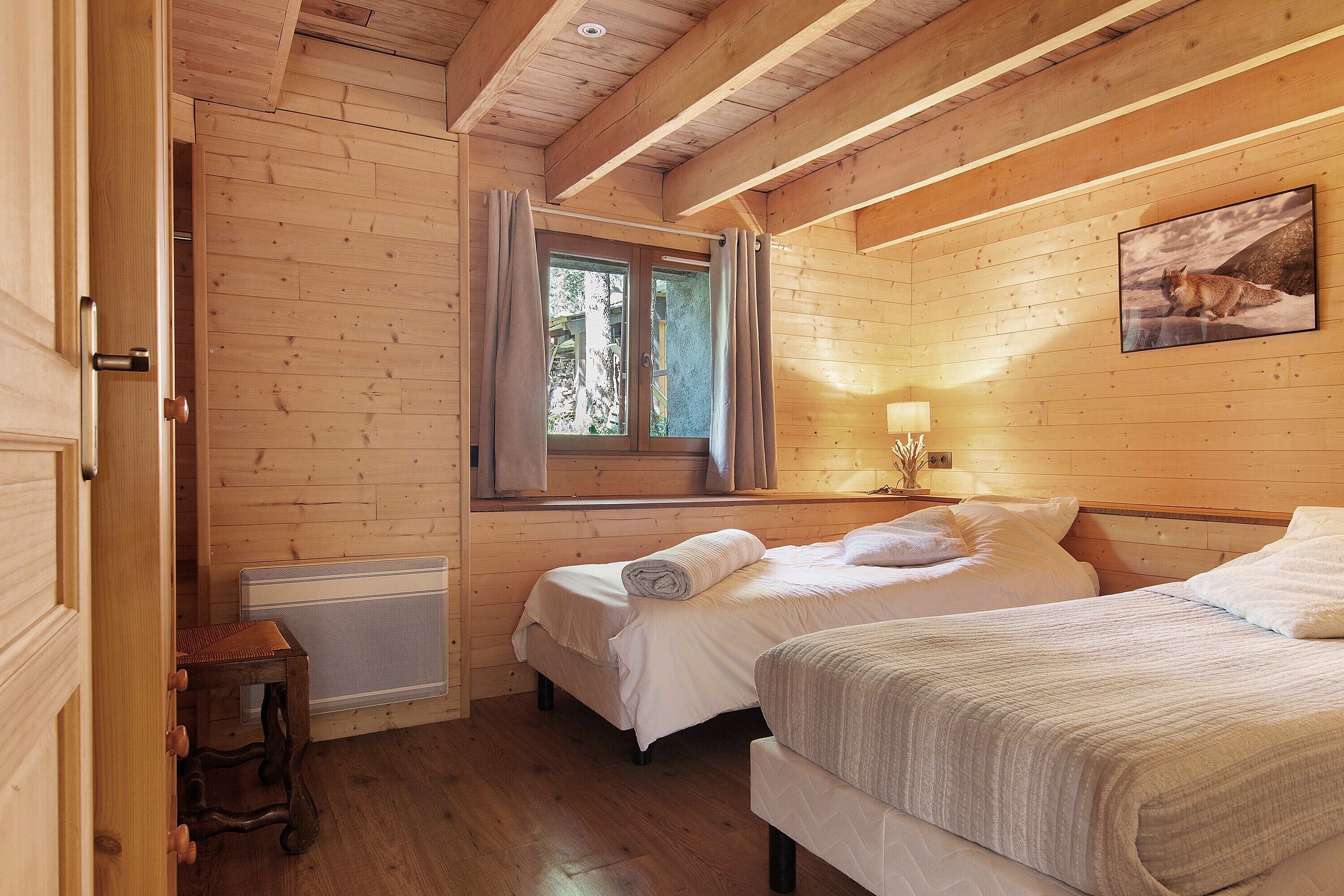 A wooden cabin bedroom with two beds, a window with curtains, a radiator, and a painting on the wall.