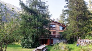 Een houten chalet met twee verdiepingen en een balkon, omgeven door dicht bos en bergen op de achtergrond.