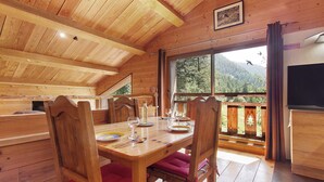 Interior de madera con una mesa de comedor preparada para dos personas, sillas y un ventanal que ofrece vistas al exterior.
