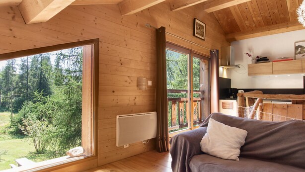 Interior acogedor de una cabaña con piso de madera, un sofá con una manta, una ventana que ofrece vista a los árboles y una zona de cocina.