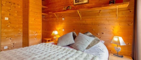 A wooden cabin room with a bed, two bedside lamps, a shelf with books, and a framed picture on the wall.