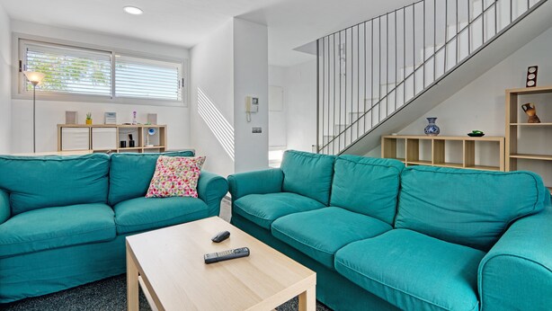 A modern living room with teal sofas, a wooden coffee table, a staircase, and a bookshelf.