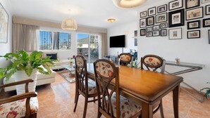 A dining area with a wooden table and chairs, a TV mounted on the wall, and a balcony with a view. - Vacation Home 'Vitamina Village' with Sea View, Shared Pool, Garden & Wi-Fi (Adeje)