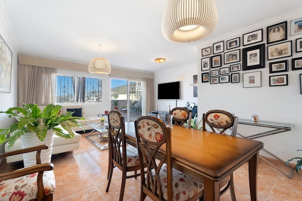 A dining area with a wooden table and chairs, a TV mounted on the wall, and a balcony with a view.