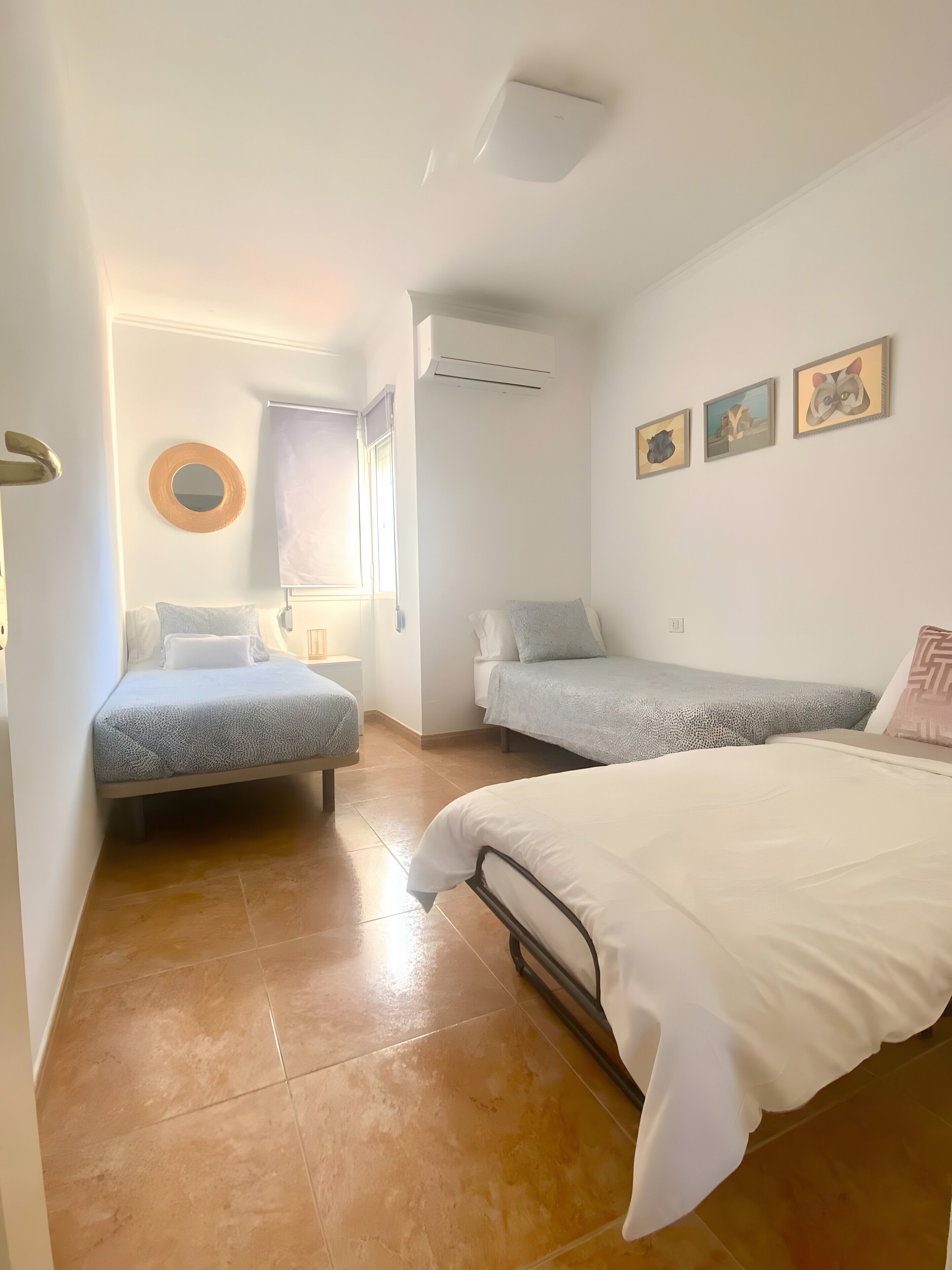A bedroom with two beds, a window with blinds, an air conditioning unit, and three framed pictures on the wall.