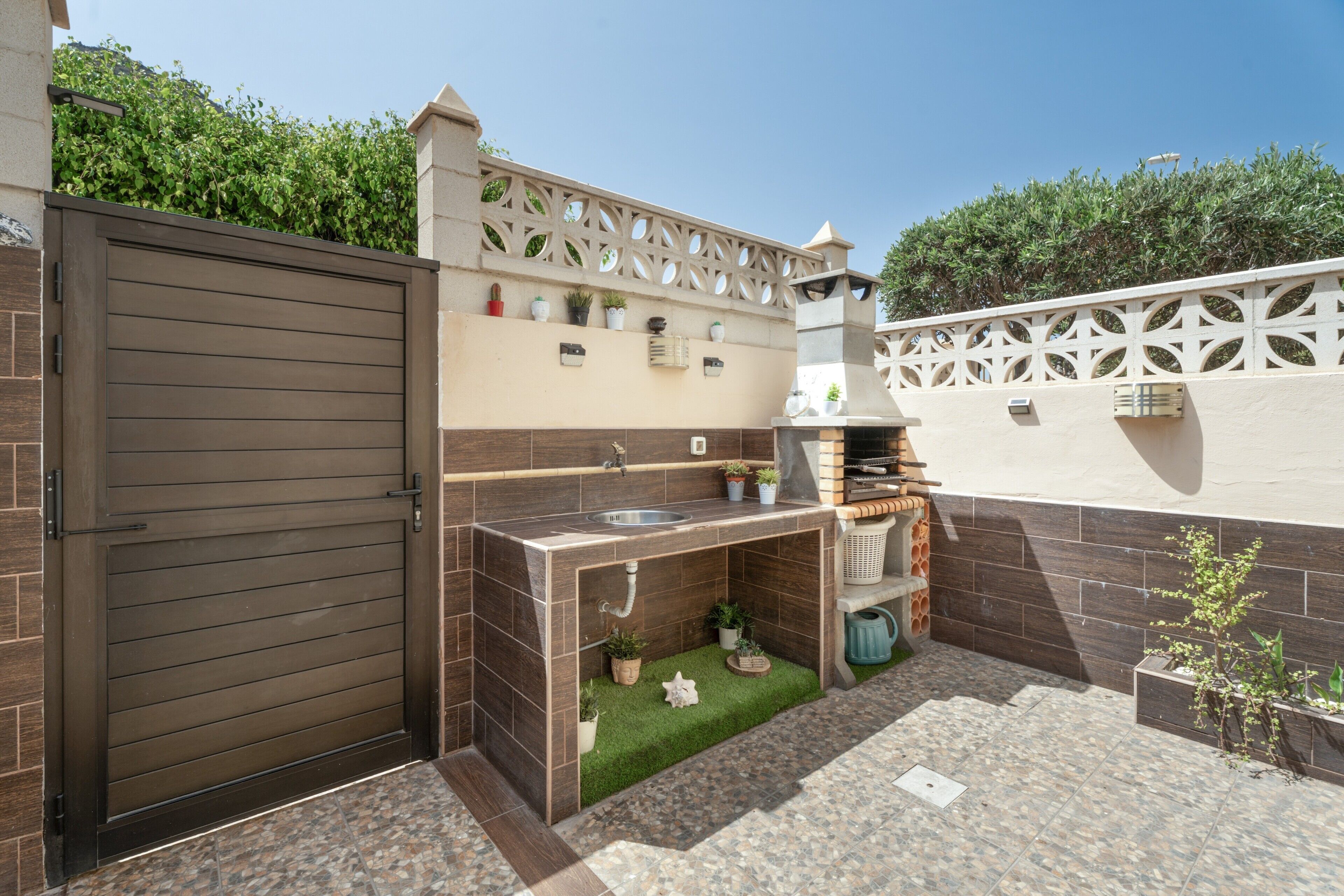 Un patio con una parrilla integrada, un lavadero y un jardín pequeño con césped artificial y plantas en macetas.