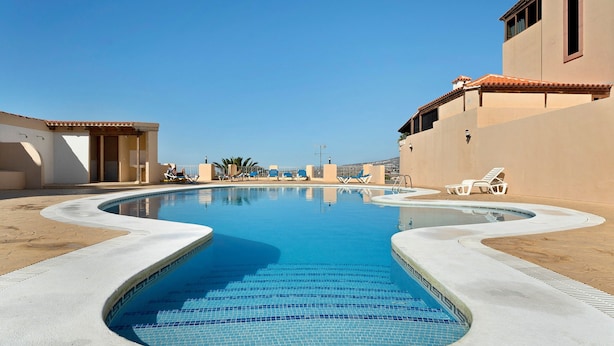 A swimming pool with a curved design, surrounded by lounge chairs and a building with a tiled roof. - Vacation Home 'Vitamina Village' with Sea View, Shared Pool, Garden & Wi-Fi (Adeje)