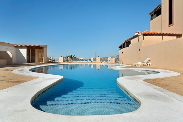 A swimming pool with a curved design, surrounded by lounge chairs and a building with a tiled roof.