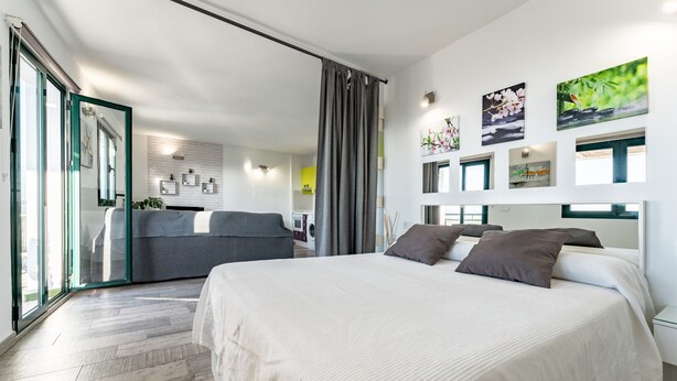 A modern bedroom with a large bed, a sofa, a dining area, and a kitchen in the background.