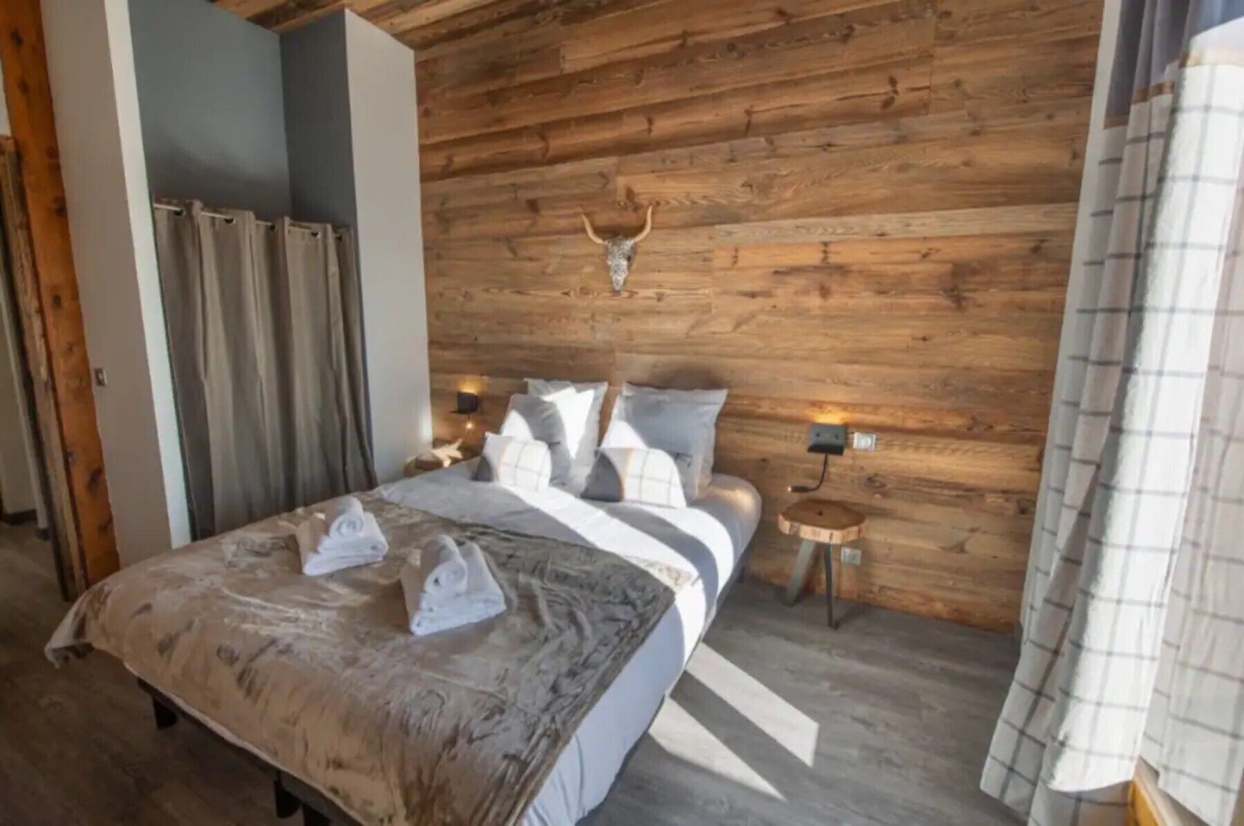 Een slaapkamer met een houten wand, een bed met witte beddengoed, twee nachtkastjes met lampen, en een hertenhoofd aan de muur.