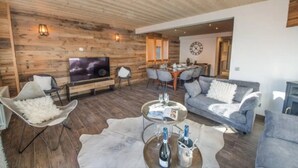 Ein modernes Wohnzimmer mit Holzbalkendecke, einem Flachbildfernseher, einem Essbereich mit Holztisch und -stühlen sowie einem grauen Sofa.