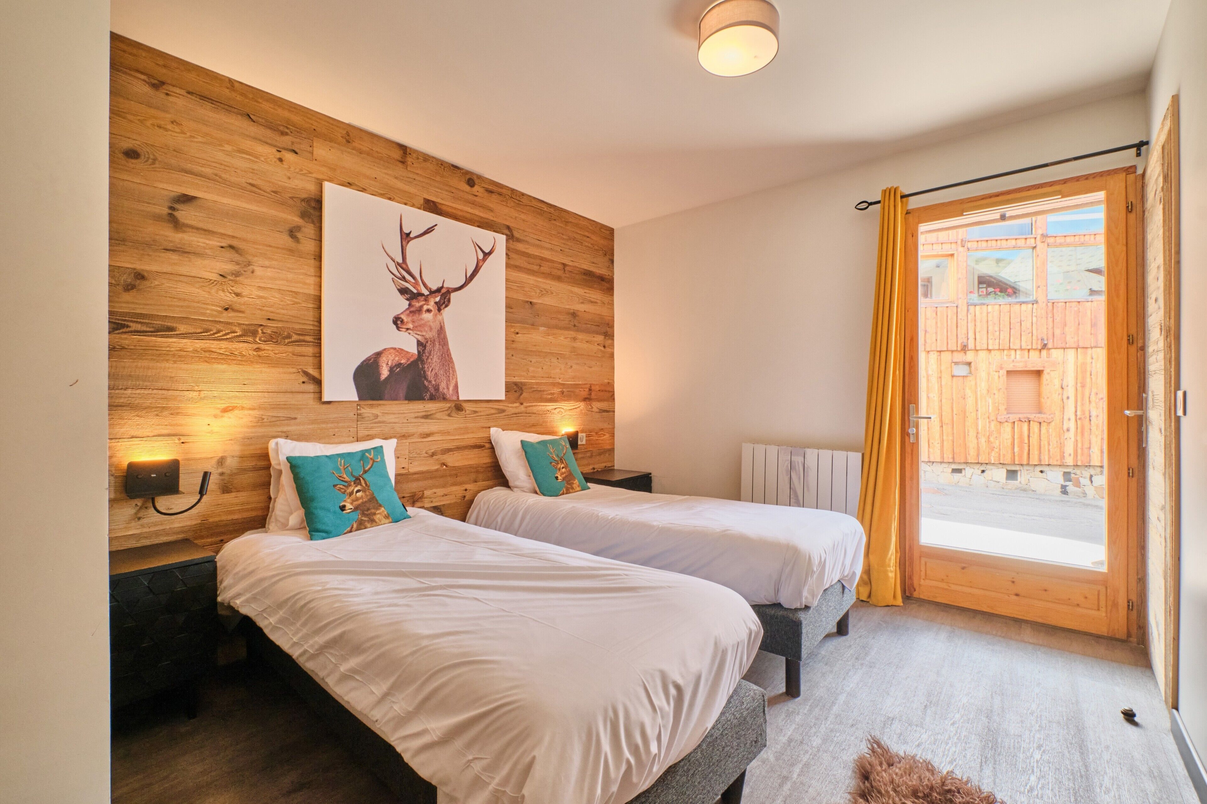 A room with two beds, wooden wall paneling, a deer artwork, and a view through a door.