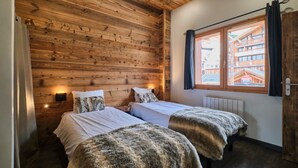 Une pièce aux murs en bois, avec deux lits recouverts de draps blancs, une fenêtre avec des rideaux et un radiateur.