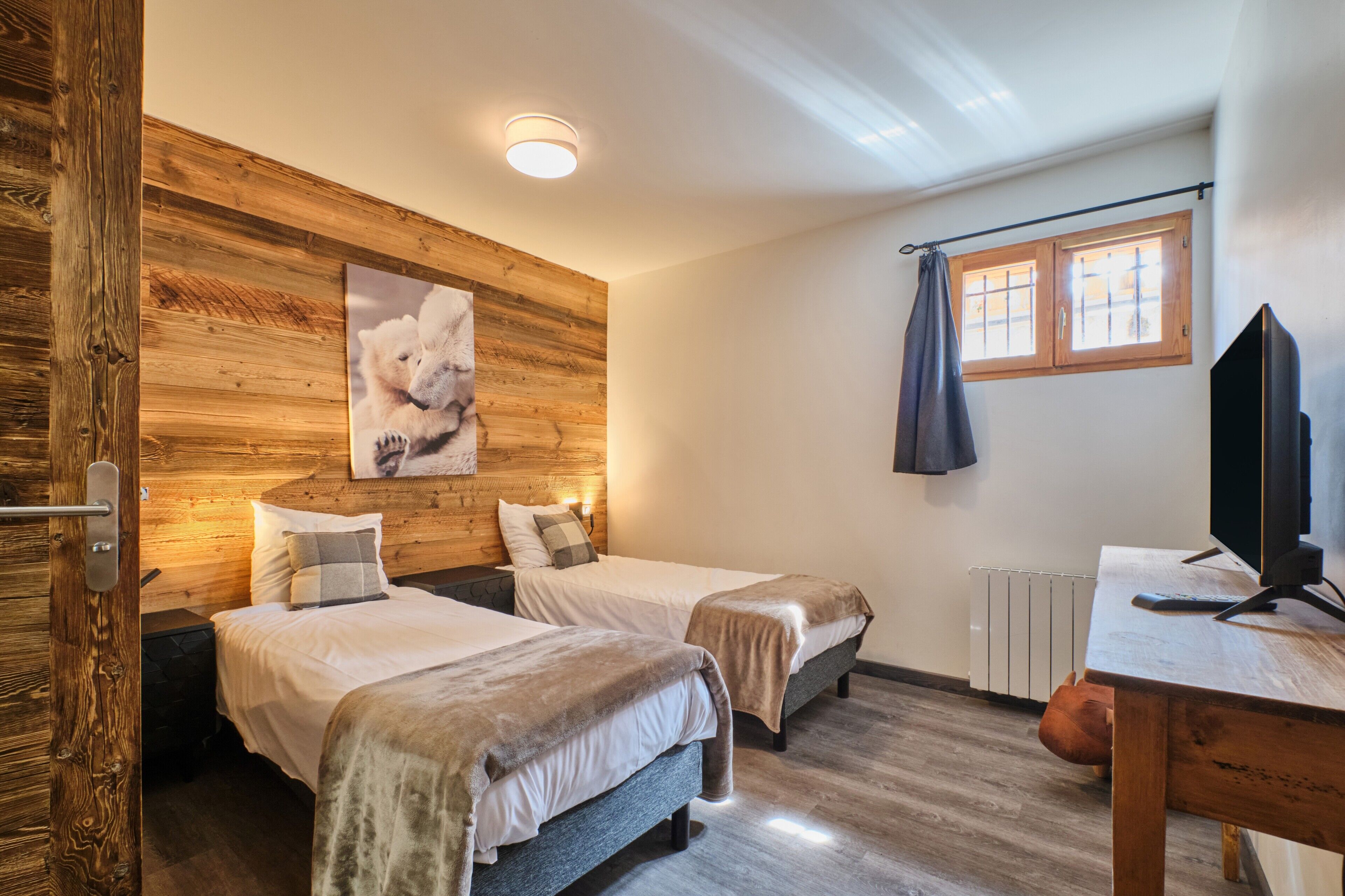 Ein Zimmer mit zwei Betten, einer Holzwand, einem aufgehängten Bild, einem Fenster mit Vorhängen und einem Fernseher auf einem Holztisch.