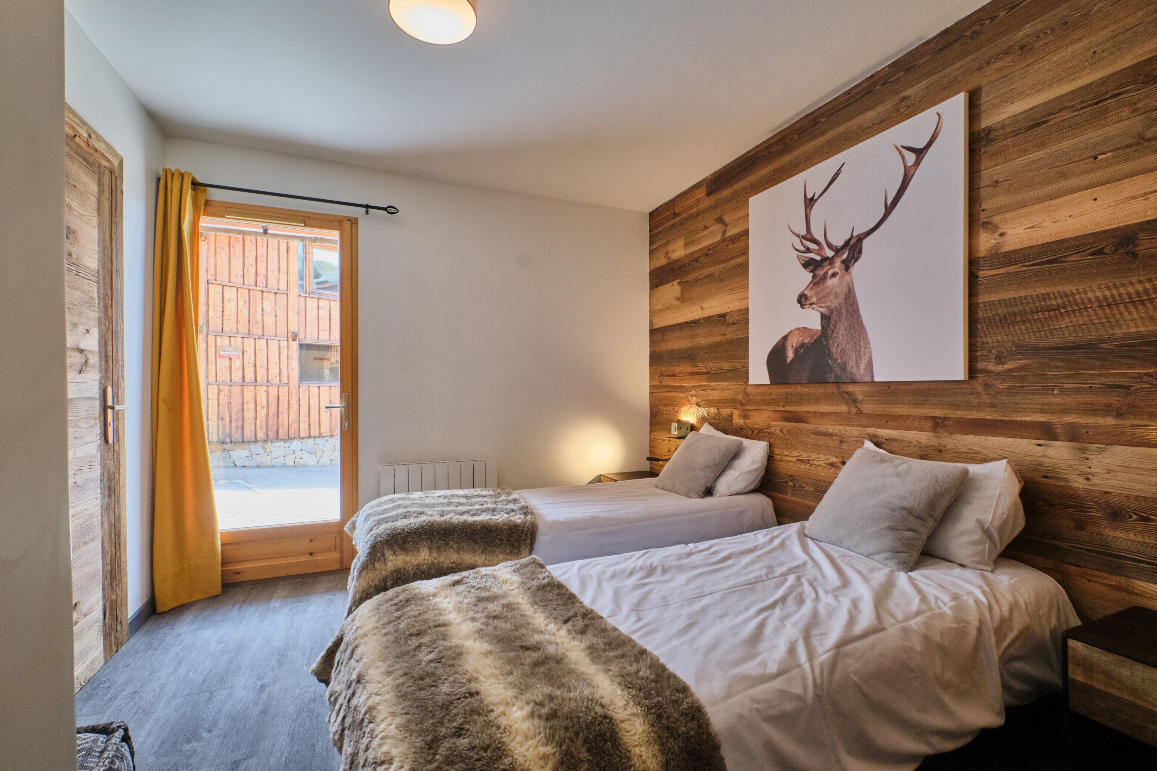 A room with two beds, a wooden wall, a deer painting, and a window with curtains.