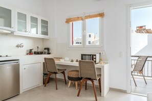 Une cuisine moderne avec des meubles blancs, un lave-vaisselle encastré, un coin repas avec deux chaises en osier et une fenêtre avec des stores.