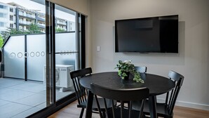 A modern dining area with a round table, black chairs, and a potted plant. A flat-screen TV is mounted on the wall above the table.