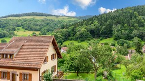 Una casa con techo inclinado, un balcón y vistas a un valle verde y colinas boscosas.
