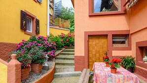 Un estrecho pasillo exterior con escalones que conducen a una puerta de madera, rodeado de plantas en macetas y una mesa con mantel colorido.