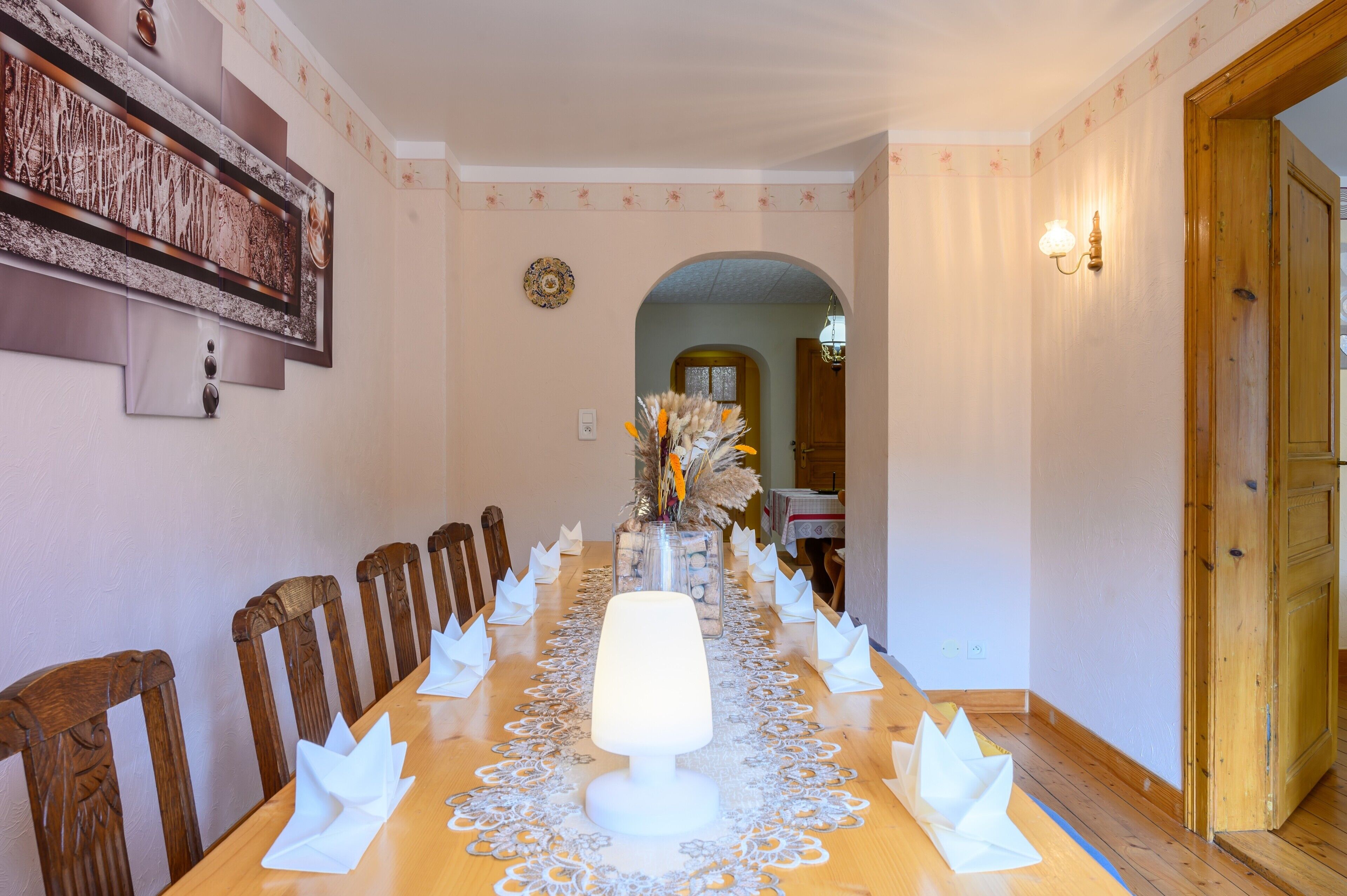 A dining room with a long table, chairs, a central light fixture, and a decorative wall piece.
