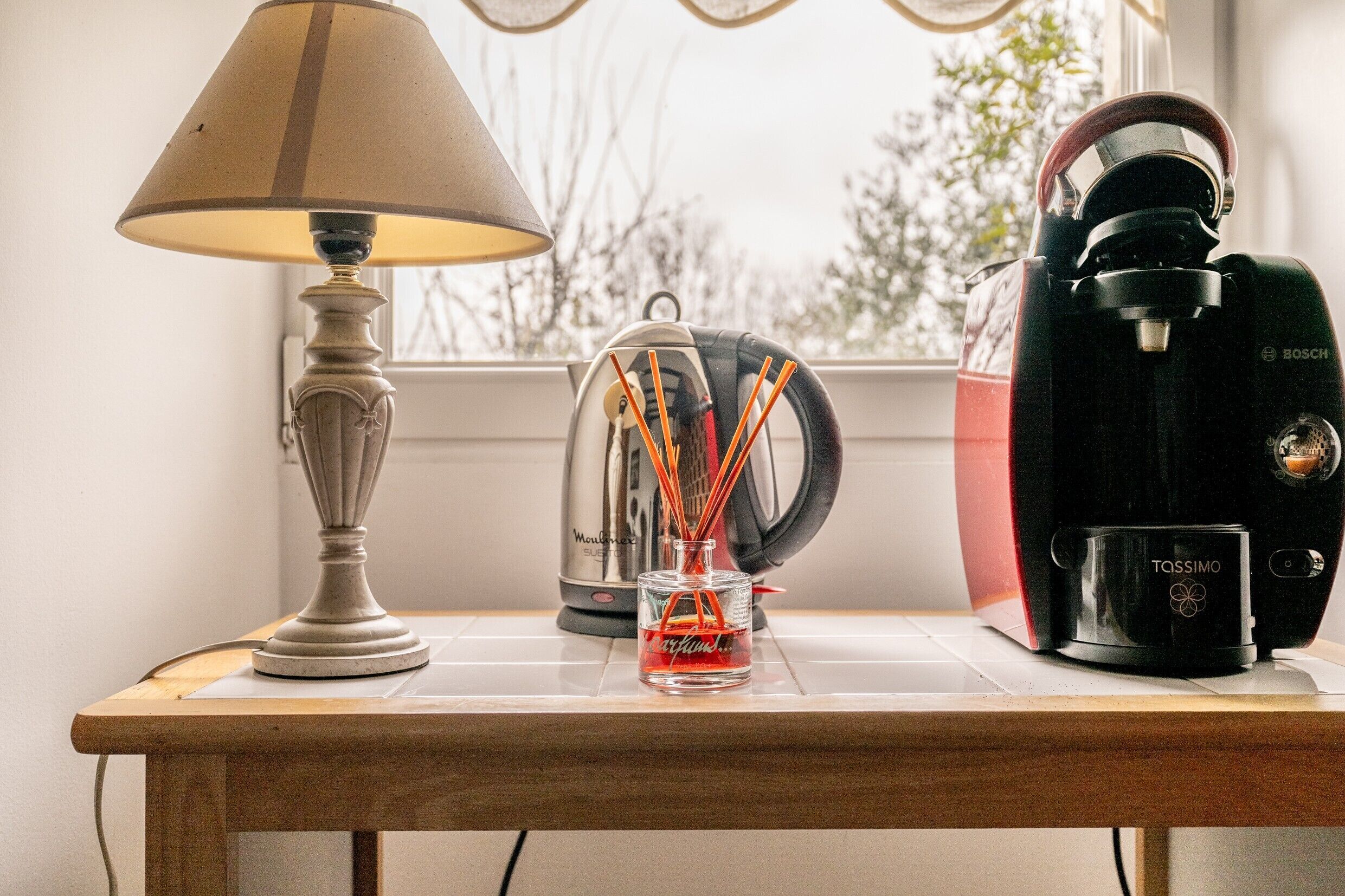 Een tafel met een lamp, een koffiezetapparaat, een glas met rode vloeistof en een waterkoker.