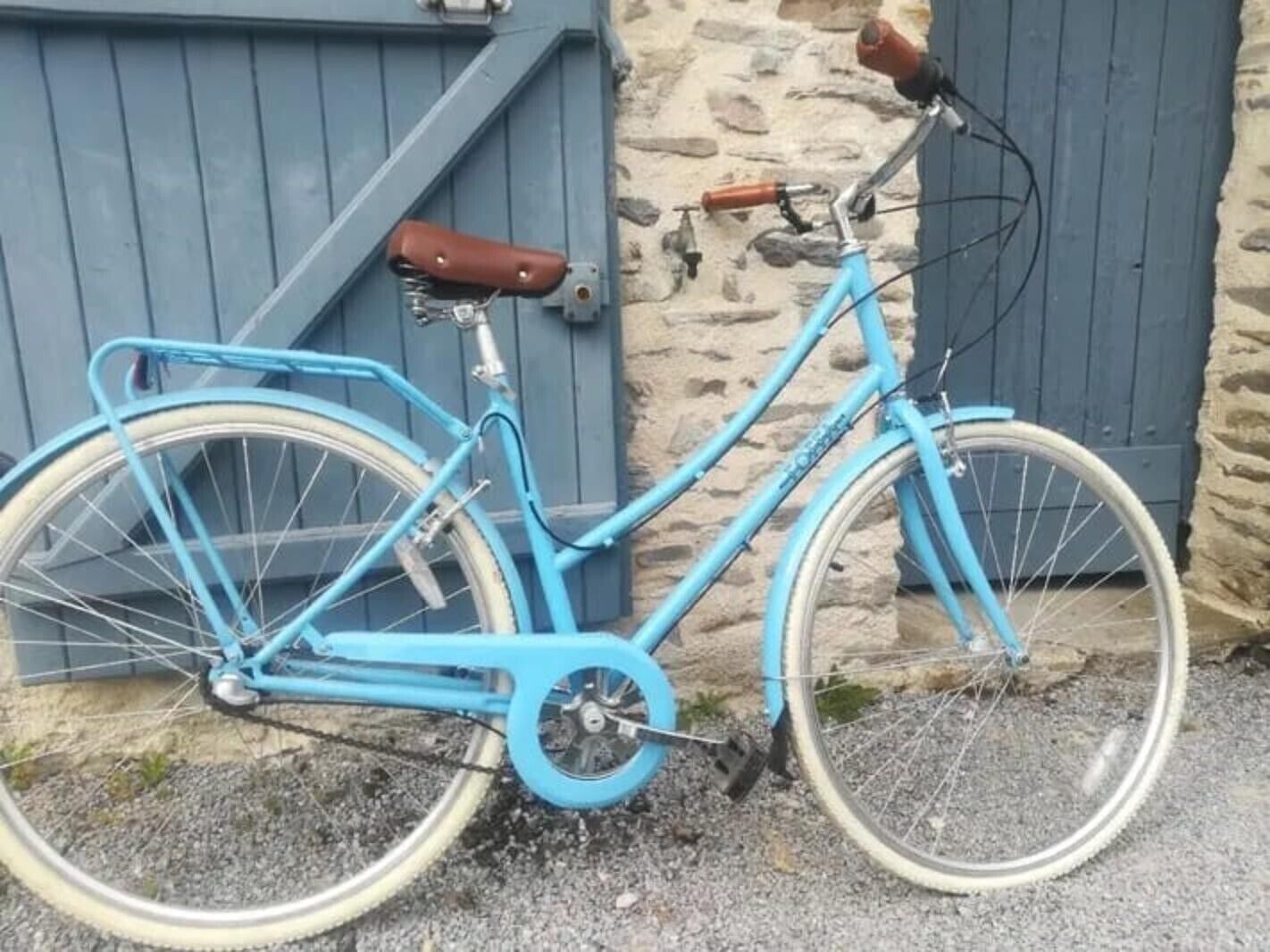 Uma bicicleta azul com selim de couro marrom e cesto frontal, estacionada encostada a uma parede de pedra e uma porta azul.