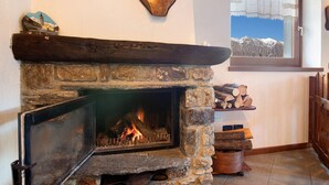 Una chimenea de piedra con fuego, una ventana con vistas a montañas, y un estante de madera con leña.