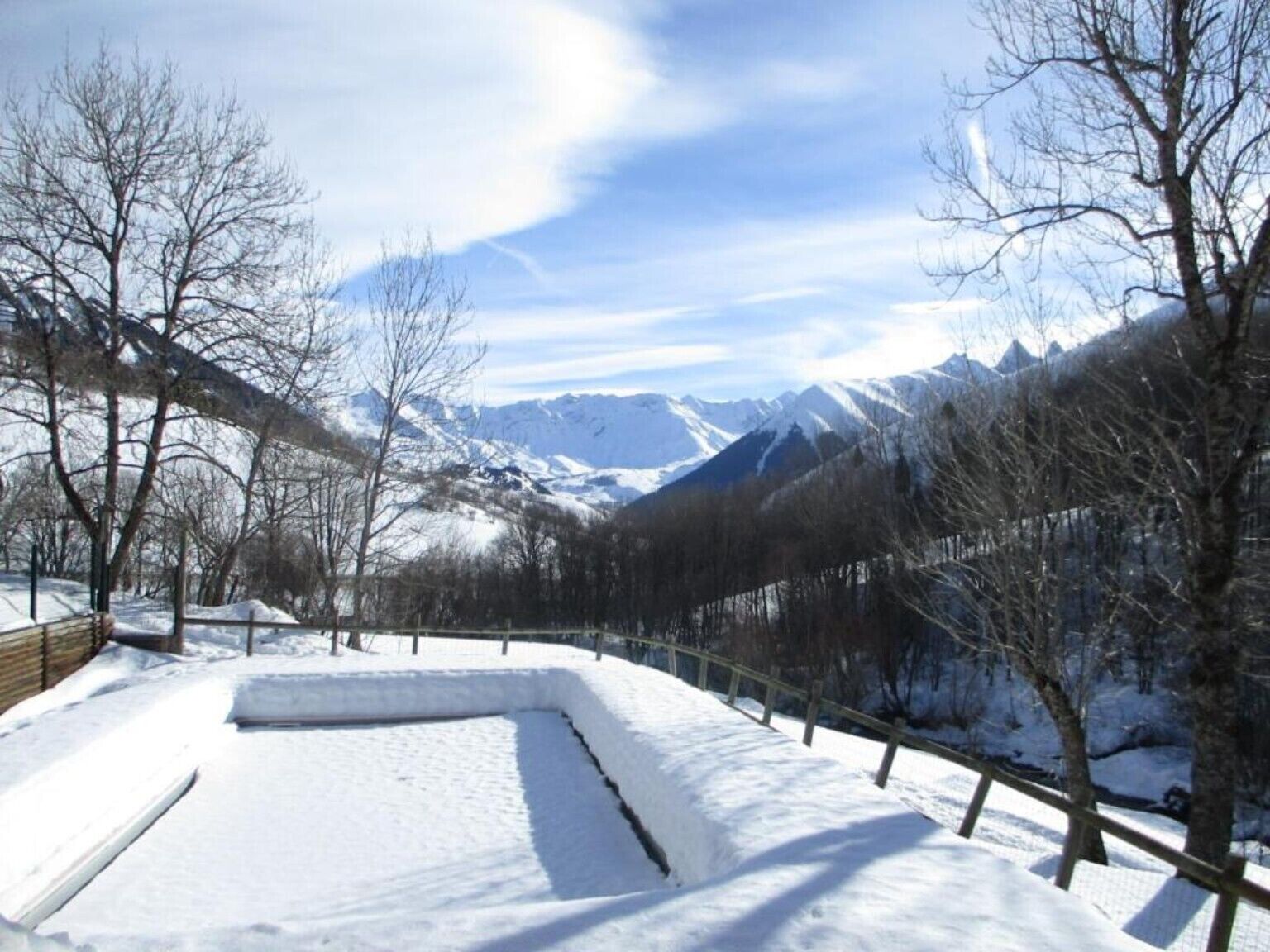 A snow-covered landscape with mountains in the background, a fenced area, and leafless trees.