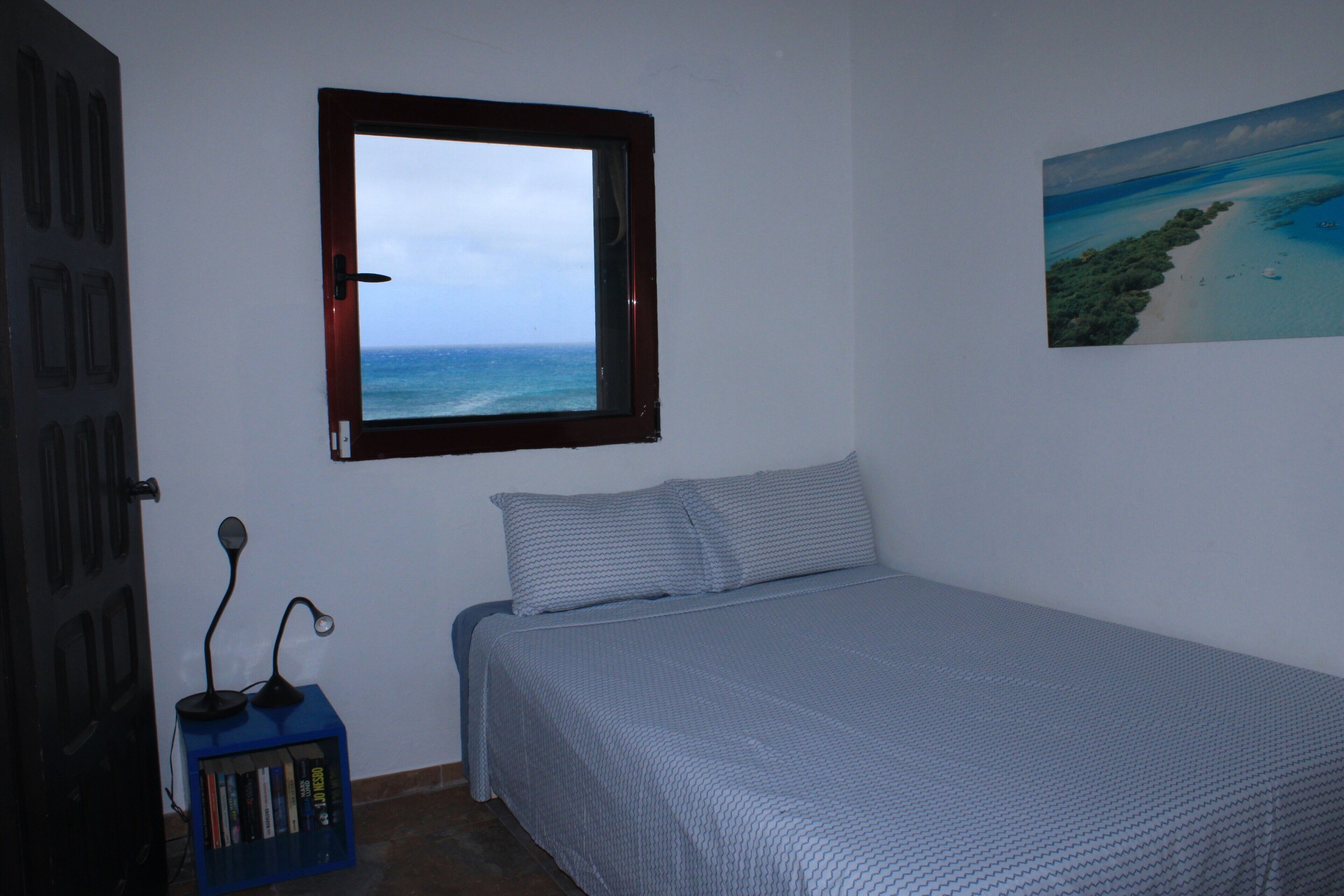 A bedroom with a bed, a window with a view of the ocean, a bookshelf, and a painting on the wall.