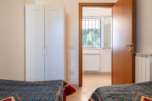 A room with two beds, a wardrobe, a radiator, and a window with a view of greenery.