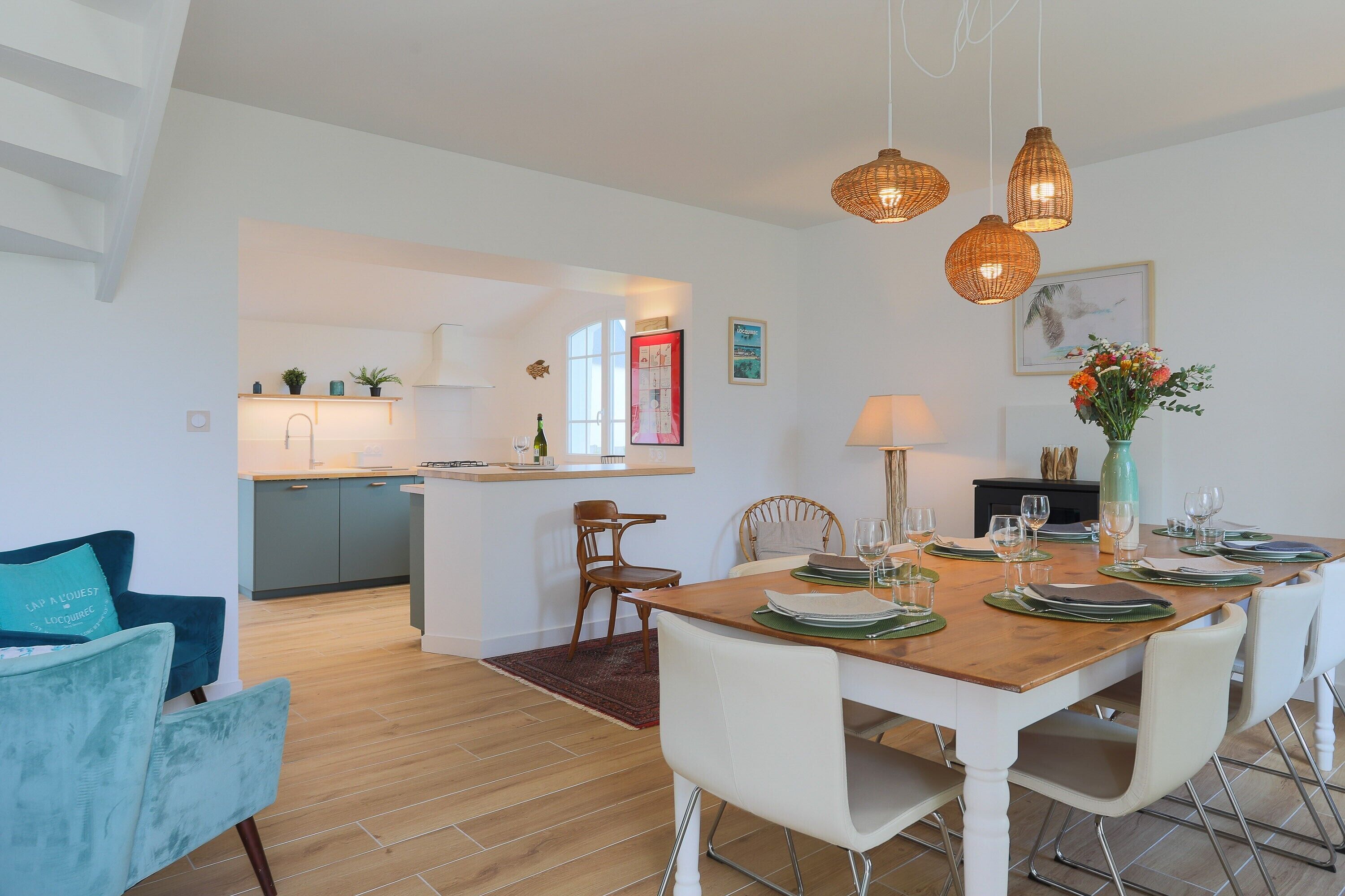 Un coin repas avec une table en bois dressée pour six personnes, des chaises blanches et une cuisine en arrière-plan.