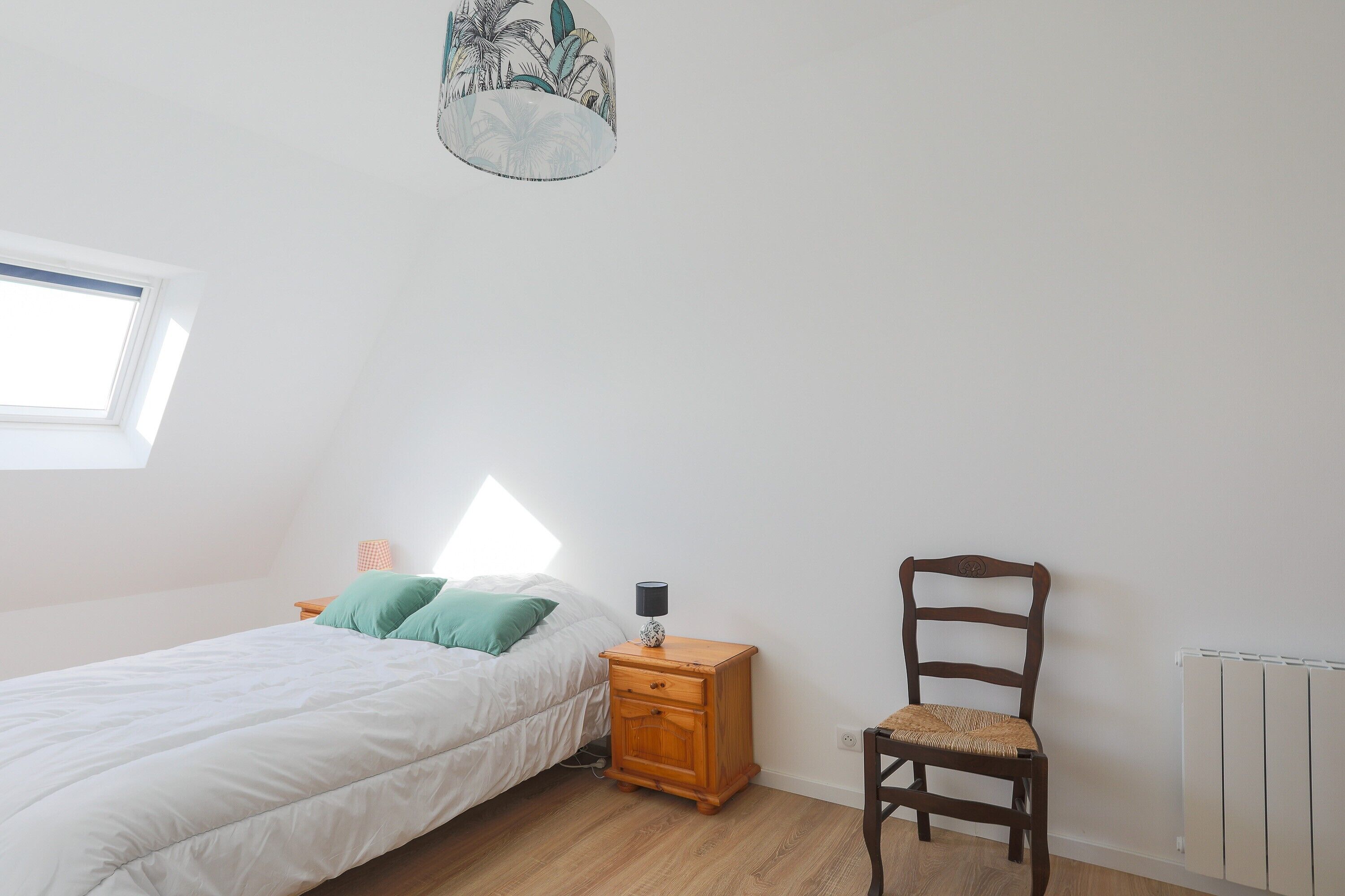 A bedroom with a bed, a nightstand with a lamp, a chair, a window, and a radiator.