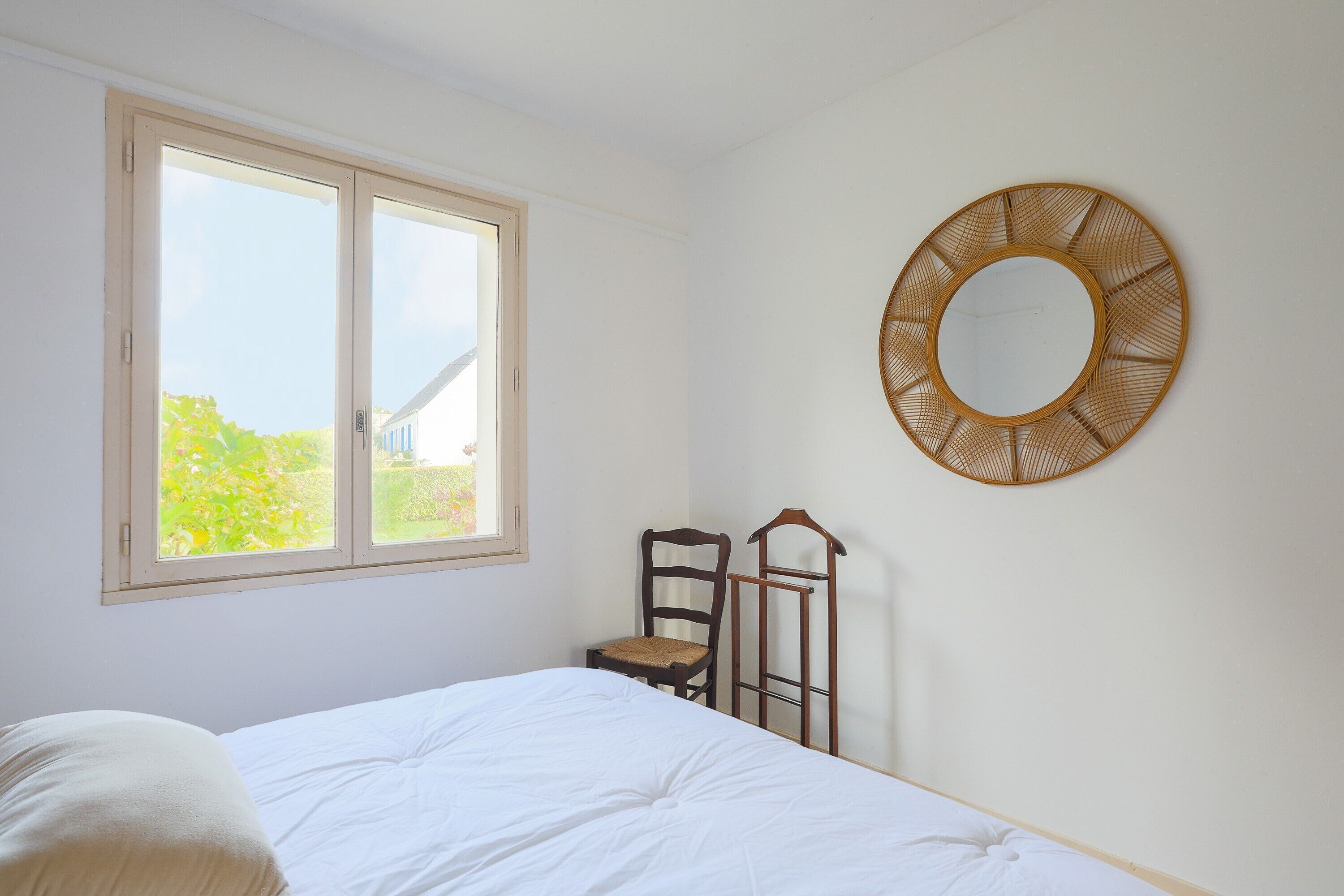 Un dormitorio con cama, ventana, una silla y un espejo fijado a la pared.